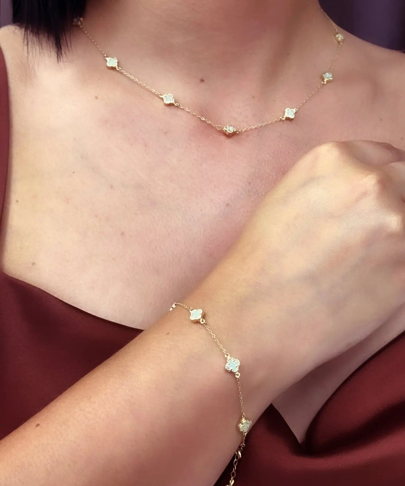 Close-up of a person wearing the Miral Jewelry 14K Yellow Gold Fashion Clover with Mother of Pearl Set, which includes a delicate necklace and bracelet, each adorned with small mother of pearl floral-shaped charms. They are dressed in a sleeveless maroon top.