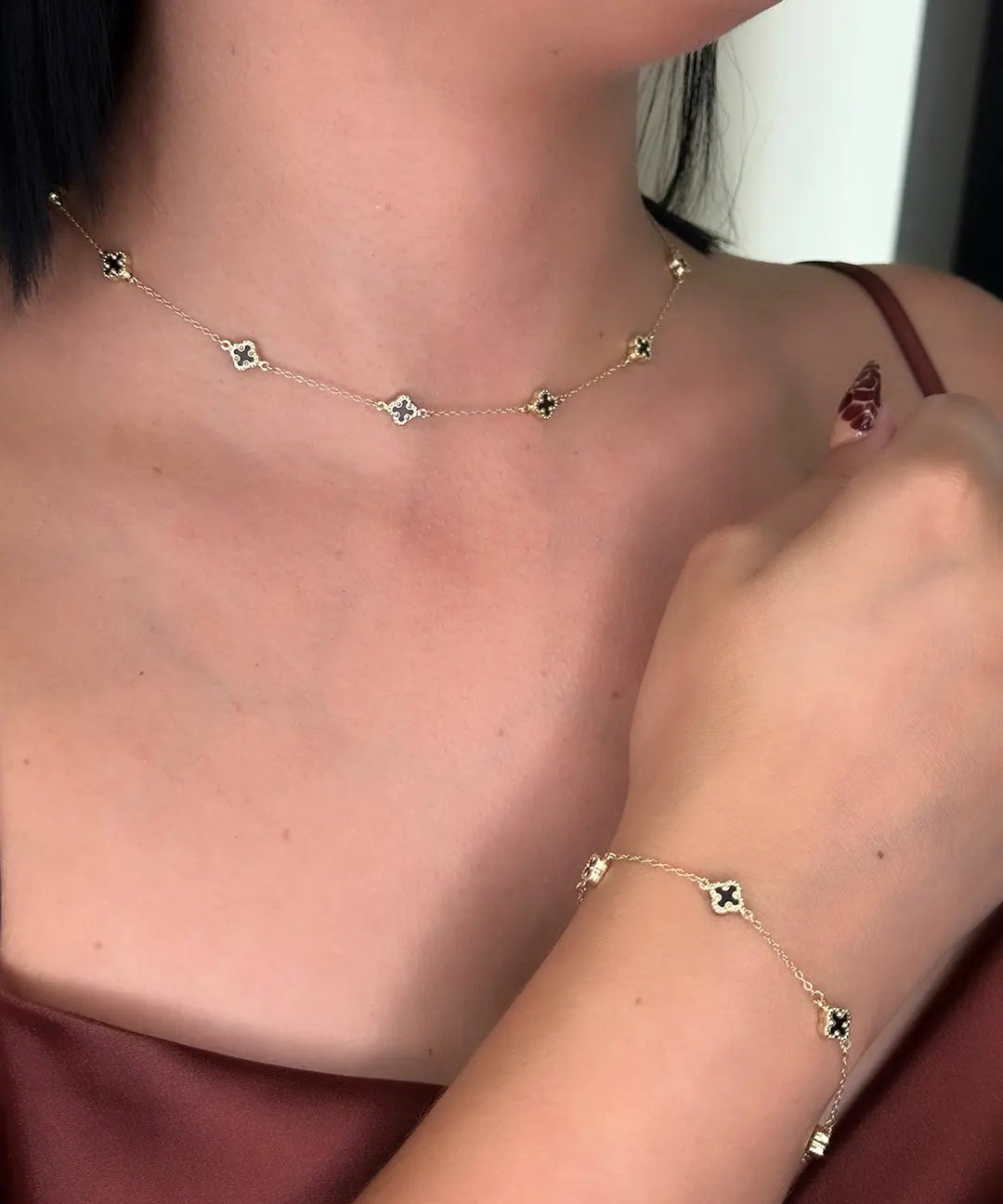 Person wearing the 14K Yellow Gold Fashion Clover with Onyx Set by Miral Jewelry, featuring a gold necklace and matching bracelet with small black clover-shaped onyx pendants, shown in close-up against a maroon outfit.