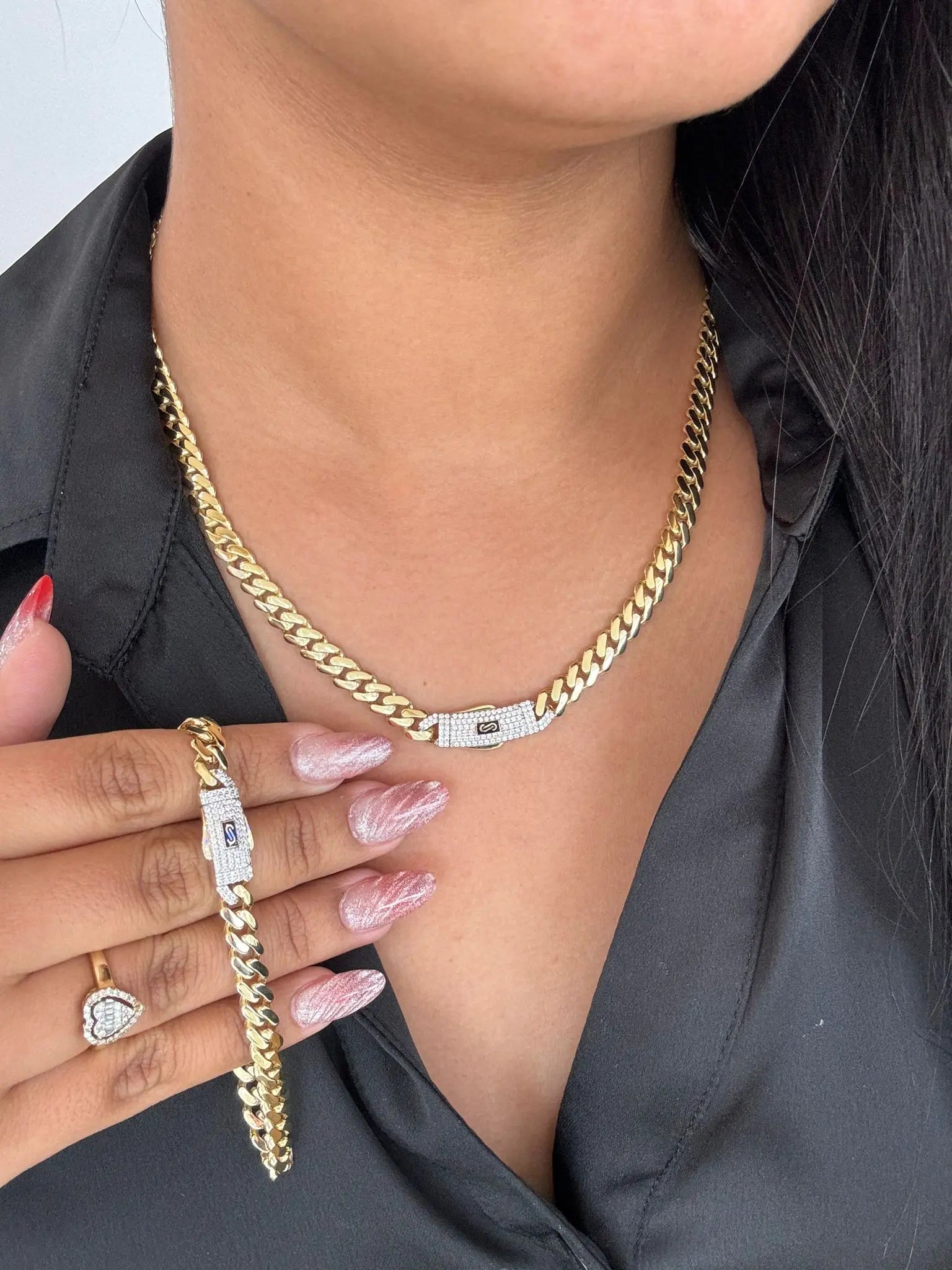 A person showcasing the Miral Jewelry's 14K Yellow Gold Monaco Set Bracelet and Chain elegantly draped over a black shirt, complemented by a matching ring with an intricate clasp design. The light pink manicured nails emphasize the exquisite jewelry ensemble.