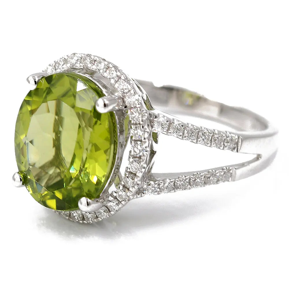 A Miral Jewelry White 14 Karat Fashion Ring Size 7 With 0.45Tw Round Diamonds And 4.88Tw Oval Peridots featuring a large round green gemstone surrounded by small round diamonds, all set in an elegant split shank design.